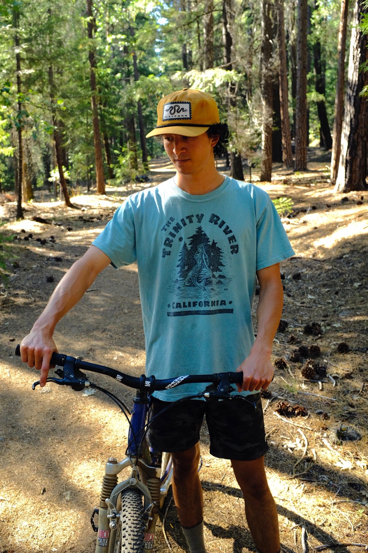 Person with a bicycle in a forest setting wearing a blue t-shirt and cap.